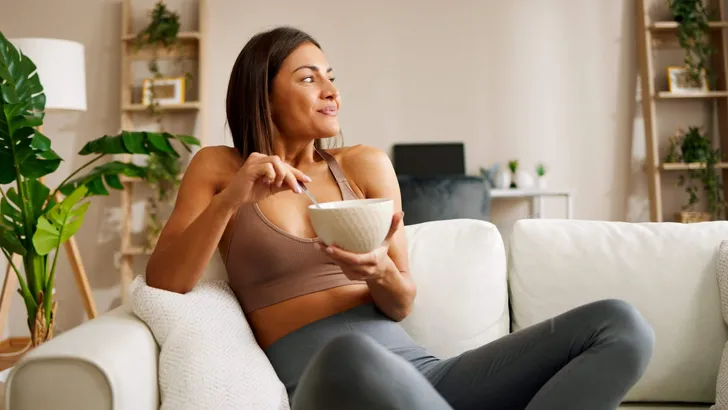 Beautiful young woman is sitting on the sofa in the living room and eating oatmeal