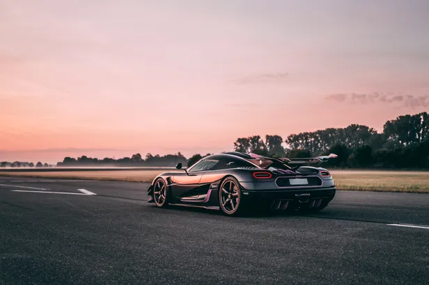 Koenigsegg One:1 in blank carbon met roze accenten, gefotografeerd van achteren op een leeg asfaltterrein bij zonsondergang.