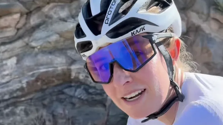 Laini Multer (Laini Bones) op de fiets in een wit fietsshirt, dragend een witte Kask Protone fietshelm en een opvallende blauwe sportzonnebril, terwijl ze lachend in de camera kijkt tegen een rotsachtige achtergrond.