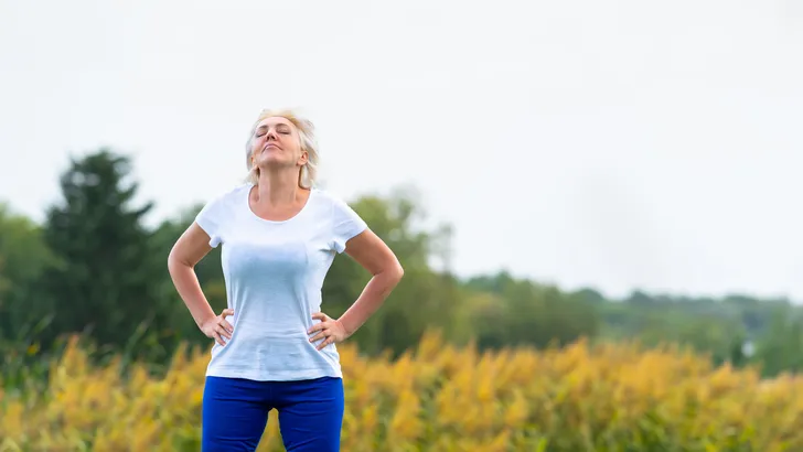 Volwassen vrouw neemt even pauze van het sporten in de buitenlucht met haar handen in de zij, kijkt omhoog.