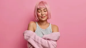 Women, tenderness, comfort. Pleased pink haired Asian woman embraces herself, closes eyes, feels coziness in soft sweater, stands against rosy background, dreams about something in peace atmosphere