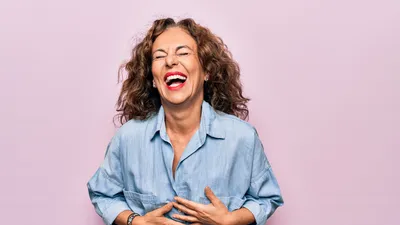 Vrouw van middelbare leeftijd staat lachend voor een roze achtergrond.