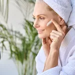 Smiling beautiful older lady looking at mirror applying cream. Skincare concept.