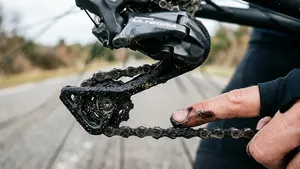 Extreme close-up van een fietsketting en derailleur bedekt met een dikke laag zwarte, zanderige oliesmurrie, waarbij een vinger wordt bevuild door de zwarte smeer.