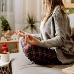 Cozy woman in knitted winter warm sweater and checkered pajama drinking hot cocoa or coffee in mug, reading book, meditation yoga on couch at home. Christmas holidays with decor and lights.