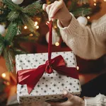 Hands in cozy sweater opening christmas gift with red bow on background of christmas tree with lights. Stylish female holding present with red ribbon in festive room close up. Merry Christmas!