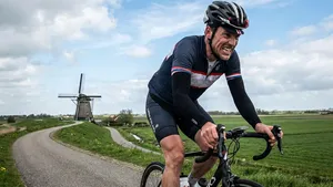 Een mannelijke wielrenner met helm en armstukken die zichtbaar afziet tijdens een zware inspanning op een dijk, met een molen in een typisch Nederlands polderlandschap op de achtergrond.