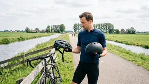 Een mannelijke wielrenner staat in een Nederlands landschap en houdt twee verschillende fietshelmen vast, alsof hij een keuze maakt tussen een goedkope en een duurdere helm.