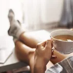 Cup of tea and chill. Woman lying on couch, holding legs on coffee table, drinking hot coffee and enjoying morning, being in dreamy and relaxed mood. Girl in oversized shirt takes break at home