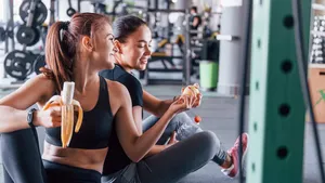 Two female friends in sportive clothes is in the gym earing fruits and taking a break