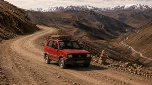 Een rode Fiat Panda 4x4 met bagage op het dak rijdt over een onverharde bergpas, met besneeuwde bergtoppen op de achtergrond.