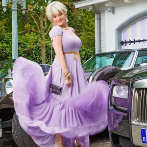 Marjan Strijbosch, bekend van The Real Housewives van het Zuiden, in een lila jurk bij verschillende luxe auto's.