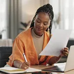 Black woman in home office, documents and laptop for research in remote work, ideas and thinking. Happy girl at desk with computer, writing notes and online search in house for freelance networking.