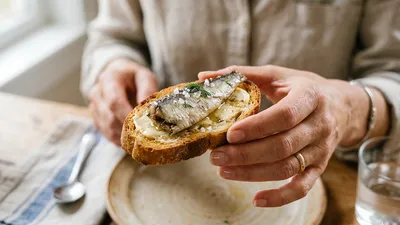 Hand van een vrouw die een rustiek toastje presenteert met een glanzend sardientje, romige boter en grof zout op een houten tafel.