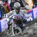 Modderige Mathieu van der Poel duwt zijn fiets door een zware crosspassage, volledig onder de modder, met publiek langs het parcours op de achtergrond.