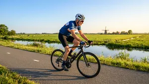 Een mannelijke wielrenner in zomerse wielerkleding fietst over een zonnige Nederlandse weg in het vroege ochtendlicht.