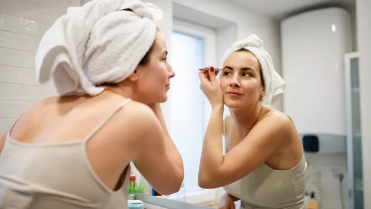 Reflection of woman with towel on hair tweezing eyebrows