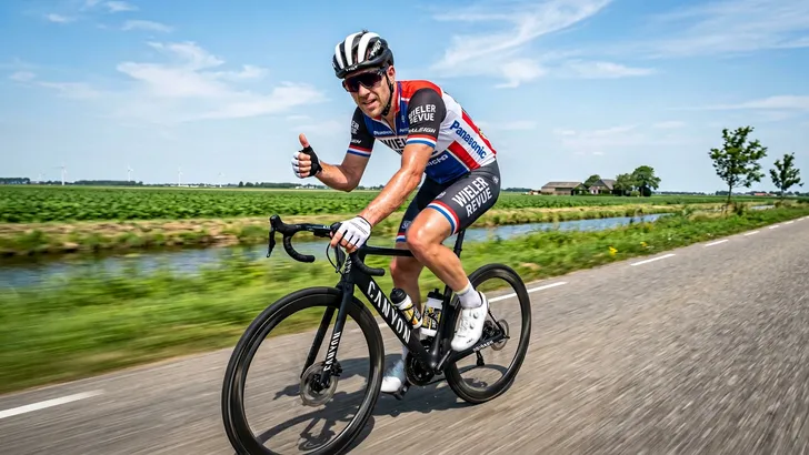 Een hevig zwetende wielrenner fietst in de brandende zon en steekt ondanks de hitte positief zijn duim op naar de camera.