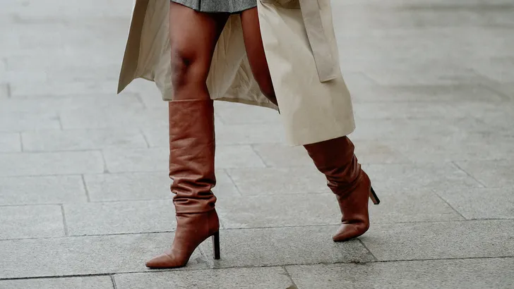 Vrouw draagt beige trenchcoat over grijze plooirok, gecombineerd met hoge bruine laarzen met hak, terwijl ze over een stenen plein in de stad loopt.