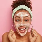 Close up shot of happy impressed curly African American woman raises clenched fists, gets pleasure from hygienic treatments, wears headband on head, washes face with bubble soap, smiles broadly