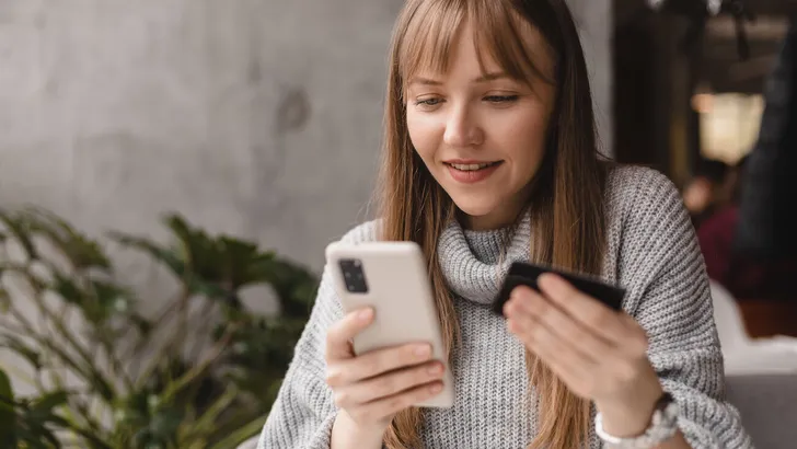 Happy young blonde woman with bang customer shopper holding credit card using cell phone mobile app buying fashion clothes paying online make purchase in ecommerce digital store on smartphone in cafe.