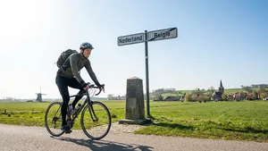 Mannelijke fietser op racefiets rijdt over de grens tussen Nederland en België met glimlach, symboliserend financiële voordelen van fietsen naar werk.