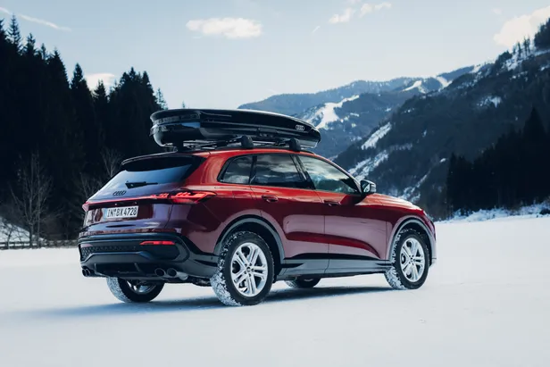Een Audi SQ5 in Grenadine Red staat geparkeerd op een besneeuwde ondergrond met bergen op de achtergrond. De auto is van achteren en van opzij te zien en is voorzien van een zwarte dakkoffer.