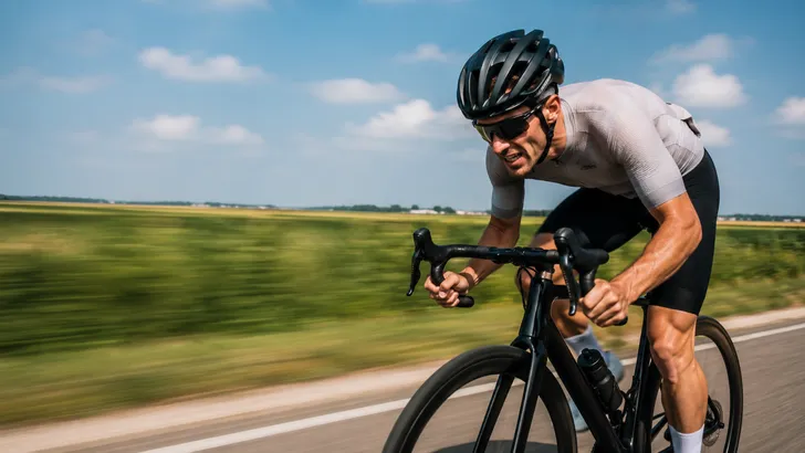 Wielrenner sprint voluit uit het zadel over een vlakke Nederlandse polderweg tijdens een krachtige inspanning, met bewegingsonscherpe groene velden op de achtergrond.