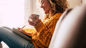 Happy cute lady at home write notes on a diary while drink a cup of tea and rest and relax taking a break. autumn colors and people enjoying home lifestyle writing messages or lists. Blonde curly beautiful lady sit down in the house