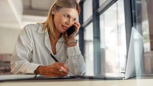 Woman entrepreneur making notes and talking by phone with client while sitting in coworking