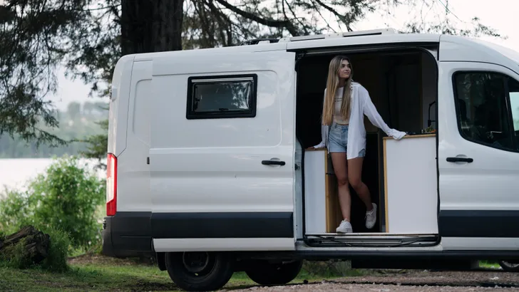Vrouw die in de deuropening van een witte camper staat en uitkijkt over het kalme meer en het omliggende bos