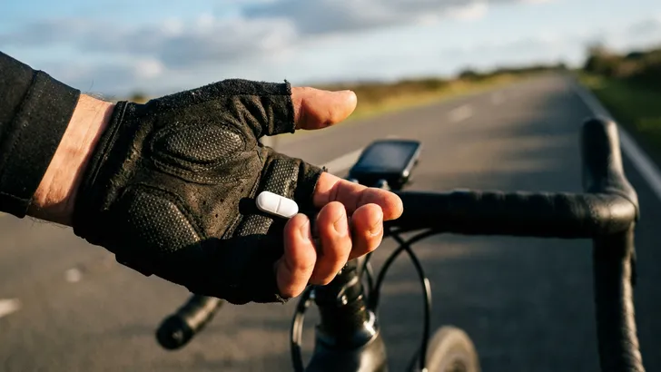 Close-up van een wielrennershand met handschoen die een witte pil vasthoudt, met op de achtergrond een onscherp racefietsstuur en een weg.