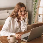 Cheerful cute beautiful business woman sit indoors in office using laptop computer listening music with earphones.