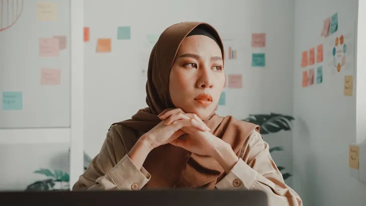 Frustrated young Asian muslim businesswoman wearing hijab working on a laptop sitting at desk in office.