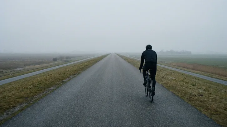 Een eenzame wielrenner rijdt op een lange, rechte en mistige dijk, wat de discipline van monotone training symboliseert.