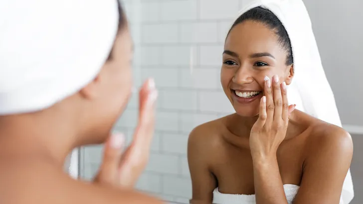 Happy woman putting on facial moisturizer with hand in bathroom mirror