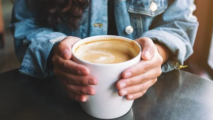 Closeup image of a woman holding a cup of hot latte coffee on the table