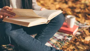 Cropped image of young woman sitting on blanket, reading book and drinking coffee or tea in autumn garden