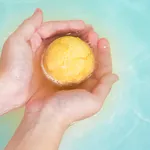 Salt bath bomb. Orange bubbling ball of bath salt in the hands on blue water background. Bomb for the bath it dissolves in the hands