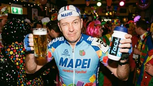 Een man in een kleurrijk wielershirt, verkleed als wielrenner voor carnaval, die lachend een groot glas bier in de ene hand en een bidon in de andere hand vasthoudt, met op de achtergrond een feestende menigte.