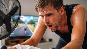 Bezwete man fietst op een indoor trainer, met handdoek op het stuur en een ventilator naast hem, Zwift-koers op tv op de achtergrond.