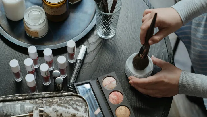 Zo lang blijft make-up houdbaar – Top-down foto van make-up op een tafel. Twee handen houden een doosje en een kwast vast.