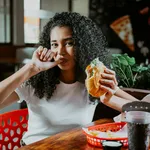 Portrait of an afro girl enjoying hamburger in a restaurant. Latin woman sucking her fingers holding a hamburger in a restaurant