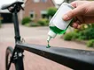 Close-up van een hand die een scheut groen afwasmiddel uit een knijpfles op het frame van een carbon racefiets spuit.