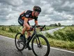 Een geconcentreerde mannelijke amateurwielrenner met fietshelm stoempt op een zwaar verzet tegen de harde wind in op een uitgestrekte Nederlandse polderweg.