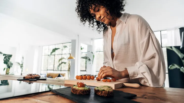 Young african american woman cutting tomatoes preparing breakfast avocado toast in bright loft kitchen.