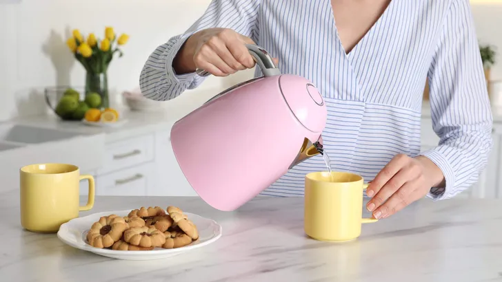 Woman pouring hot water from modern electric kettle in cup indoo