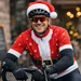 Wielrenner in rood kerstman-wielershirt met kerstmuts over zijn helm, glimlachend op de racefiets voor een café met warme kerstlichtjes op de achtergrond.