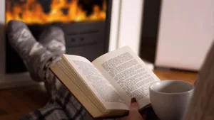 Woman reading book and hold cup of tea and warming feet in front of the fireplace