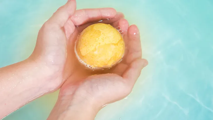 Salt bath bomb. Orange bubbling ball of bath salt in the hands on blue water background. Bomb for the bath it dissolves in the hands